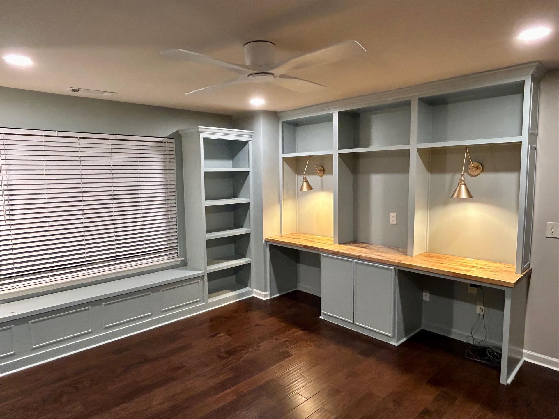 A room with a desk , shelves , a window and a ceiling fan.