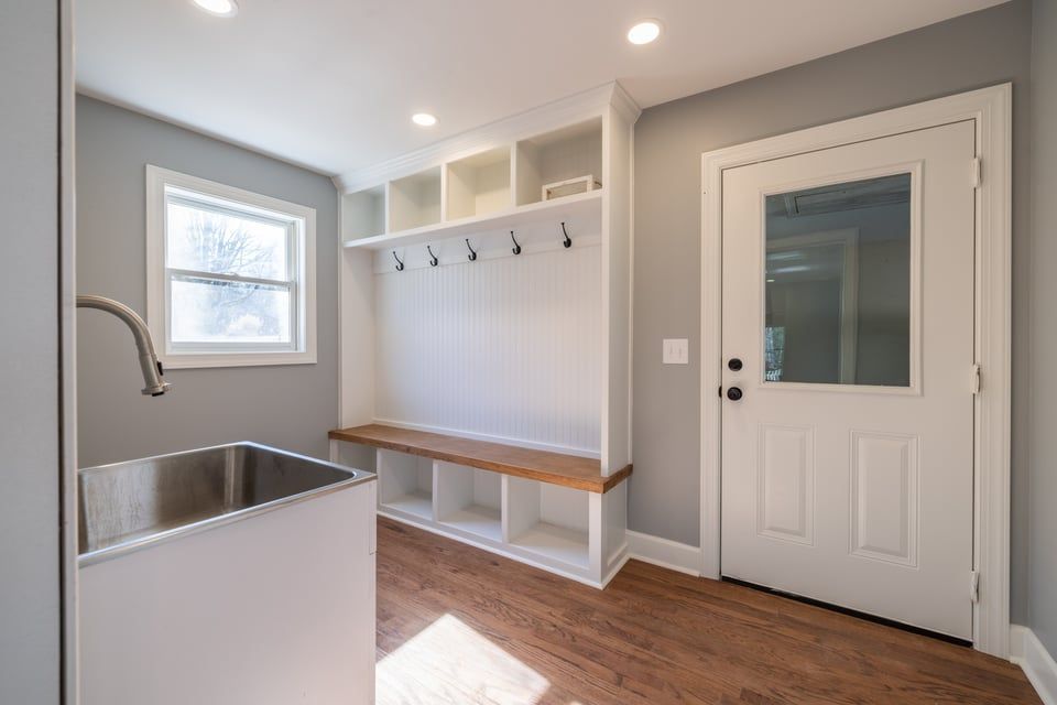 Bright mudroom with white built-in bench, hooks, cubbies, sink, and a white door on gray walls.