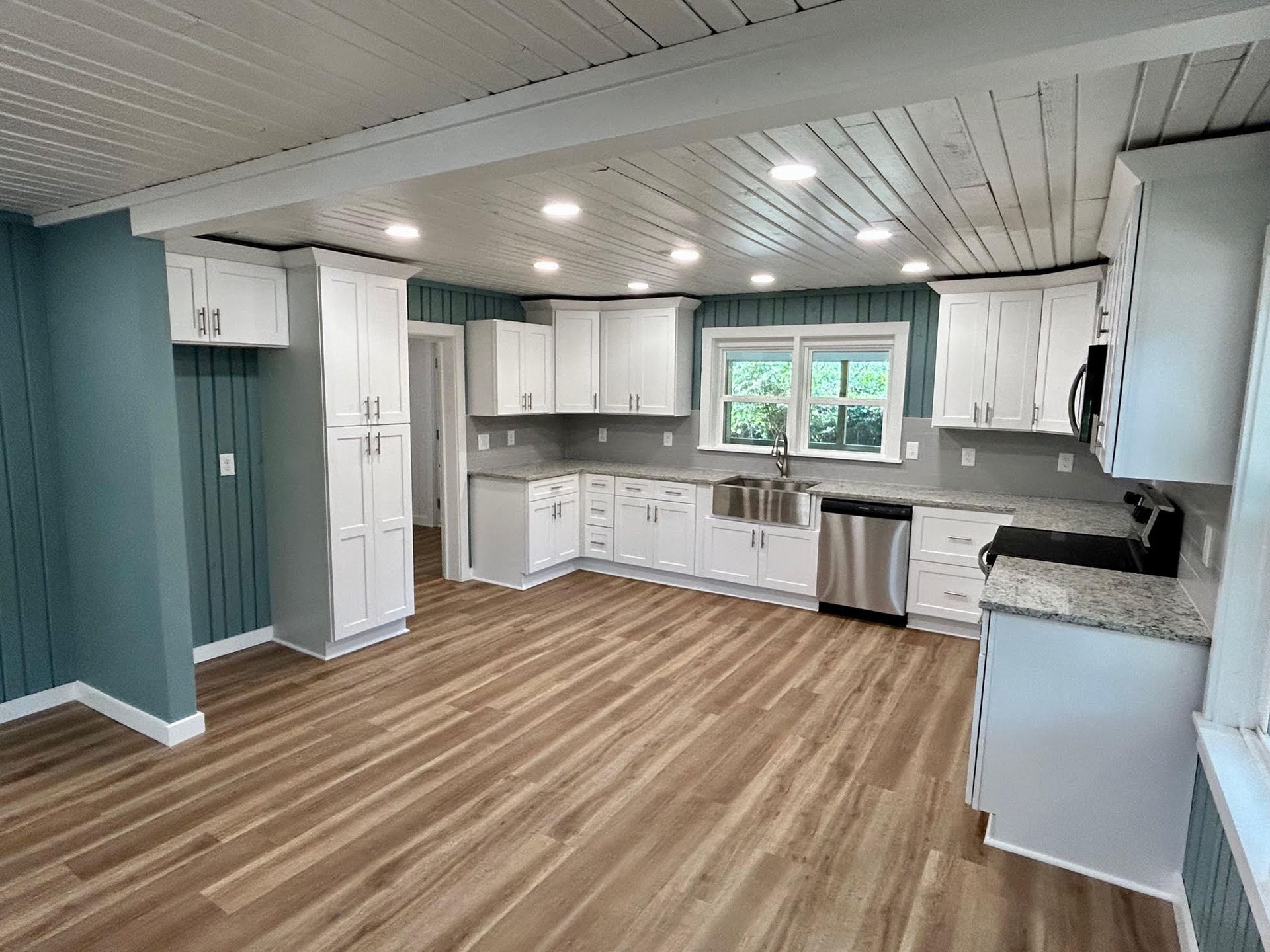 Empty modern kitchen with white cabinets, gray counters, wood floors, and a window above the sink.