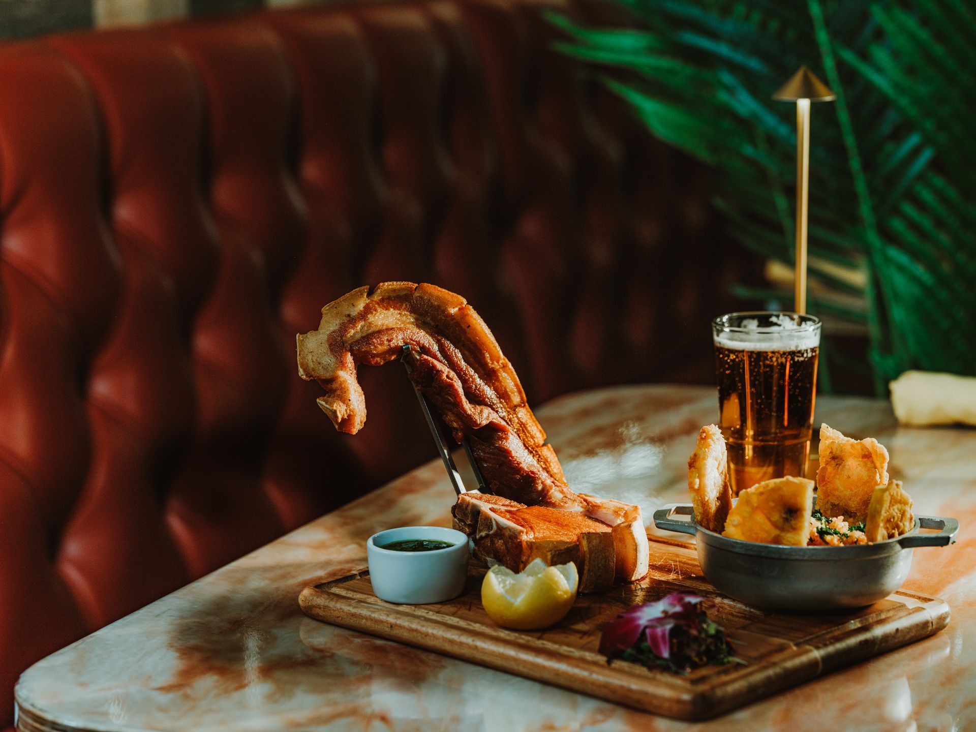 Shared platter of grilled meat and sides on a restaurant table with drinks and a tufted booth background