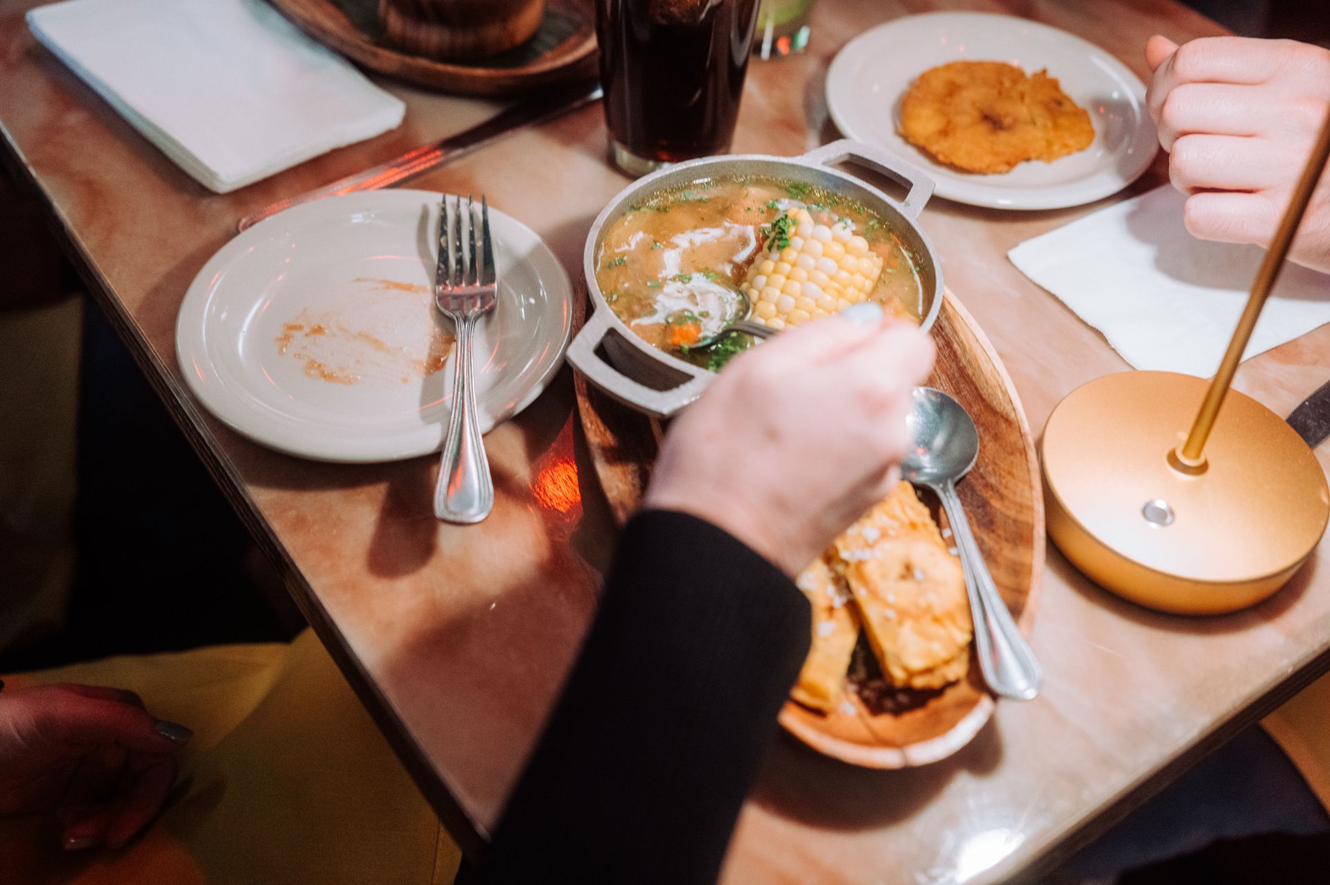 Hands reaching over a wooden table with bowls of soup, bread, and a drink during a meal