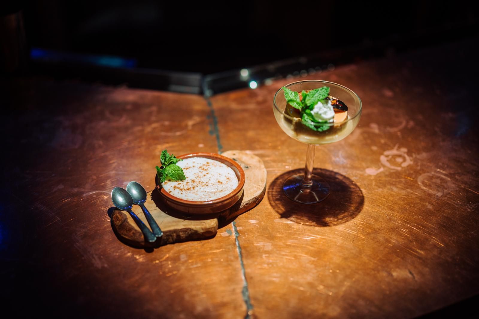Two plated desserts with mint on a wooden table, one in a glass and one in a small bowl, beside a spoon.