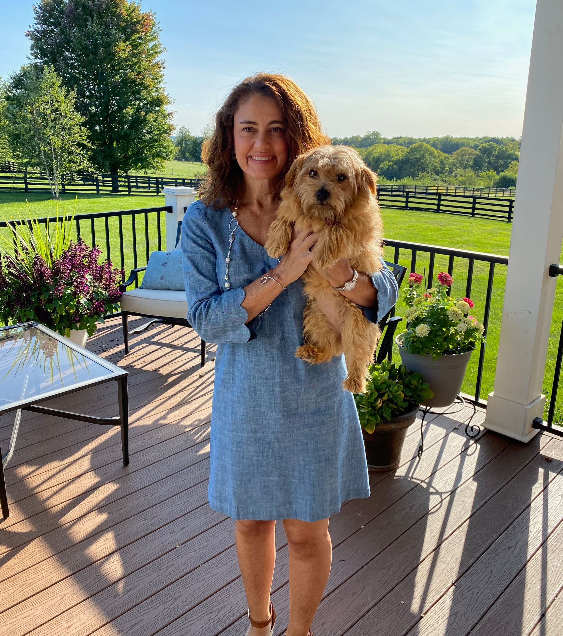 Woman in blue dress holds a small, fluffy brown dog on a deck overlooking a field.