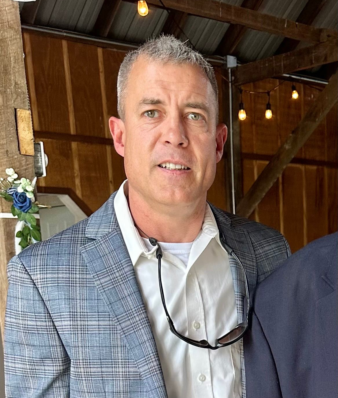 Man in plaid jacket smiles, wearing a white shirt and holding sunglasses, inside a wooden building.