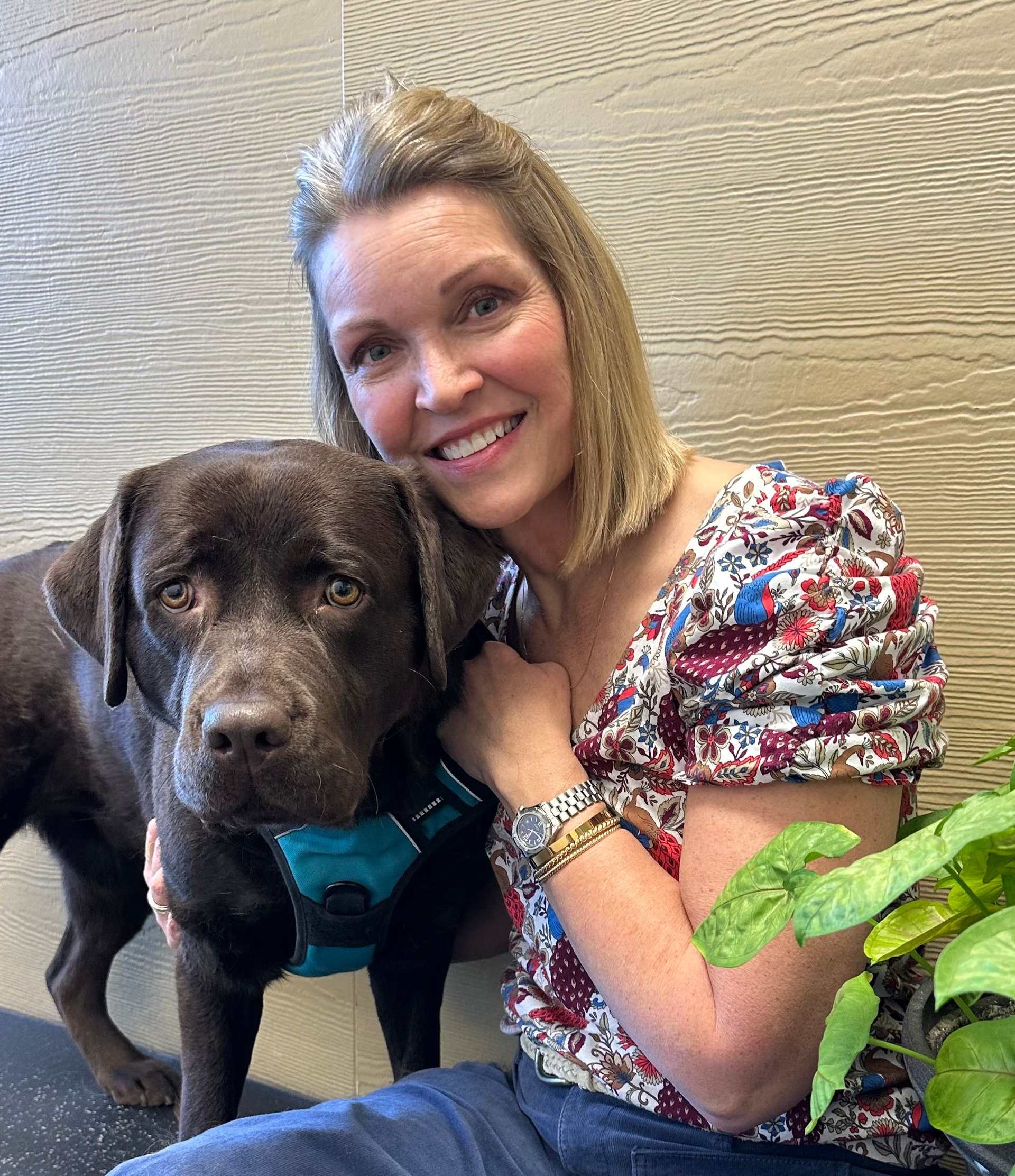 Woman with light brown hair smiles next to a brown dog wearing a blue harness.