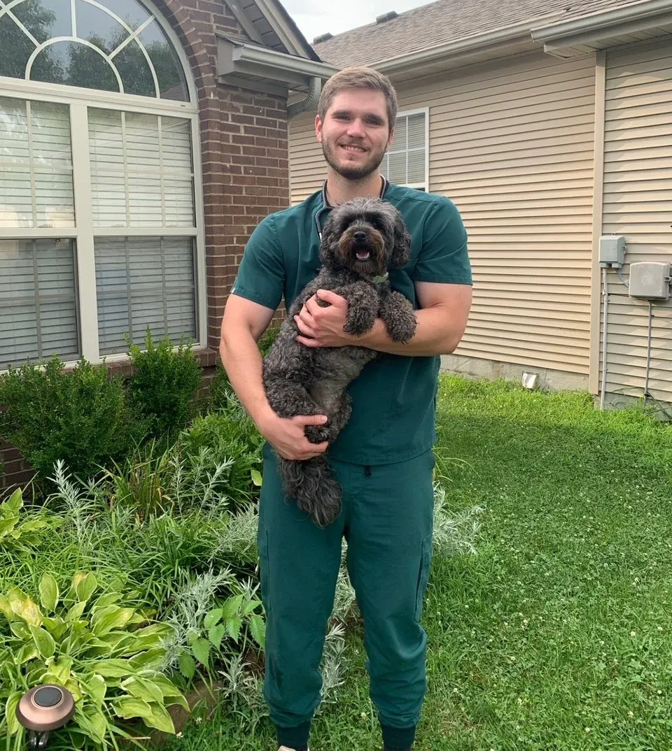 Man in green scrubs holding a gray dog outside near a house and garden.