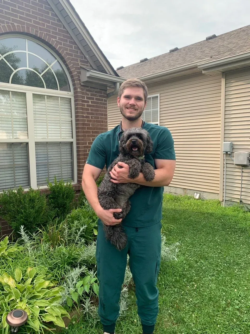 Man in green scrubs holding a small, black dog in a yard.