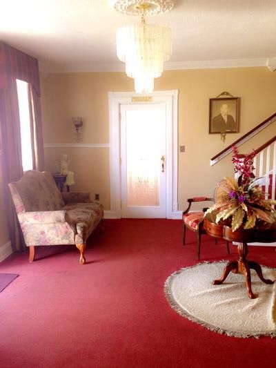 An interior view of a foyer with red carpeting, a patterned loveseat, a tiered chandelier, and a round pedestal table.
