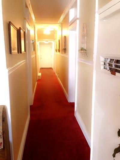 A long, narrow hallway with warm-toned walls and bright red carpeting leading to a white door at the far end.