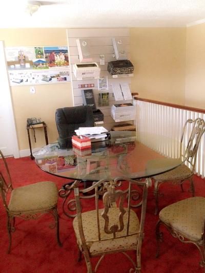 An office area with a round glass table and four chairs on red carpet, featuring wall-mounted printers and supplies.