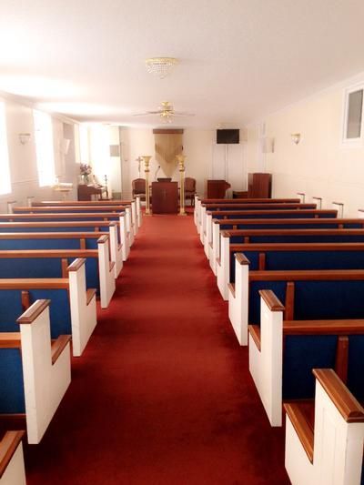 Inside a chapel with a red-carpeted aisle between rows of blue-cushioned wooden pews, facing a small altar and pulpit.