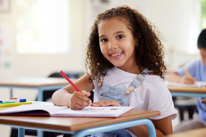 A young girl is sitting at a desk in a classroom writing in a notebook.