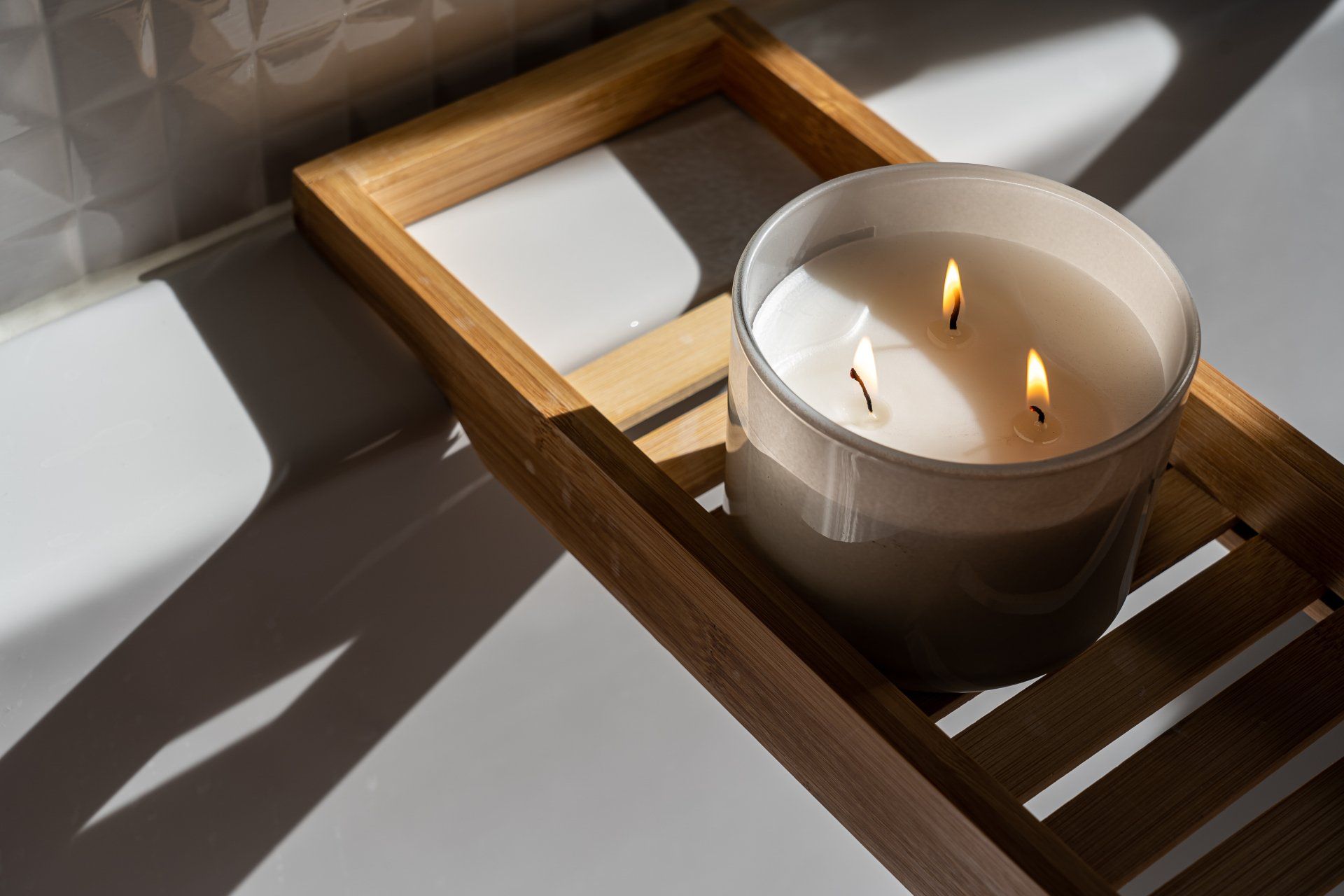 Lit candle in a glass jar on a wooden bath tray over a white bathtub.