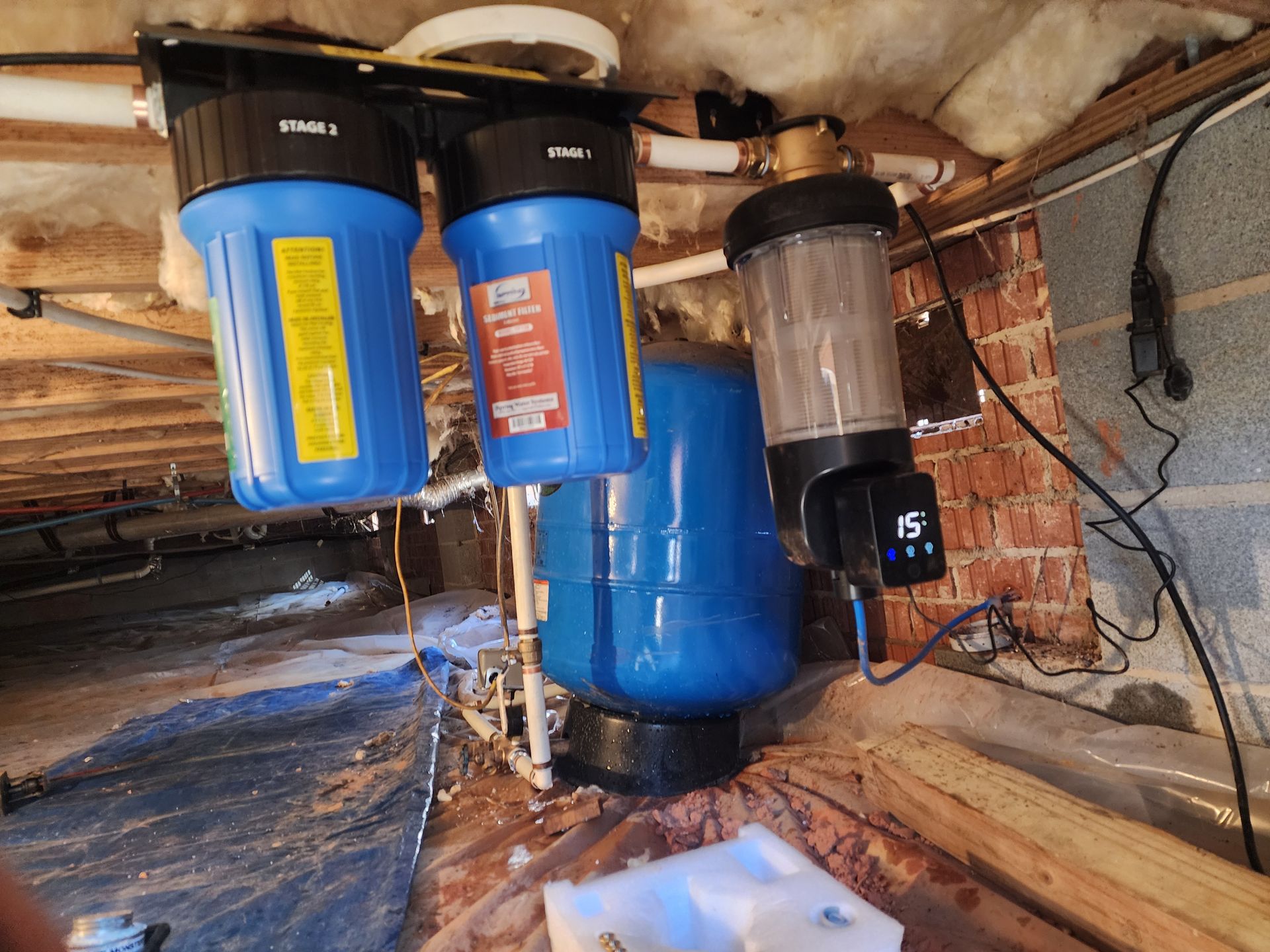 Water filtration system in a crawl space, featuring blue filter housings, a clear tank, and plumbing.