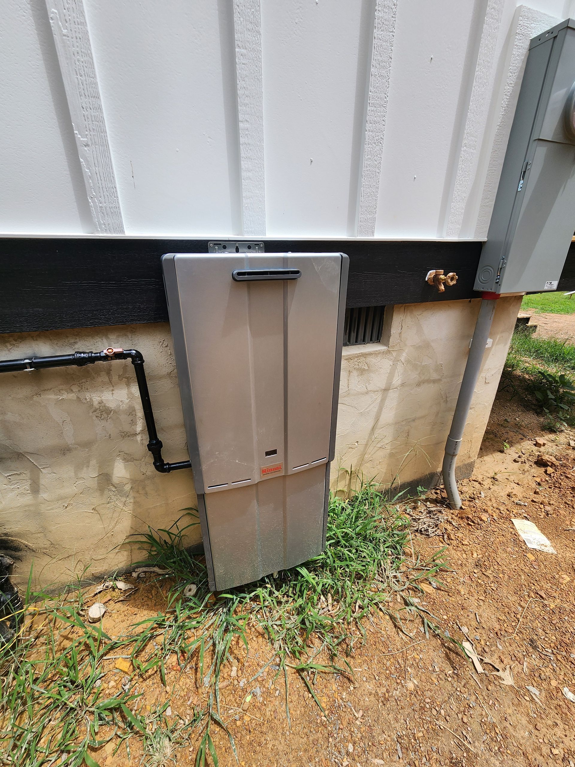 A silver tankless water heater mounted on a concrete foundation outside a house.