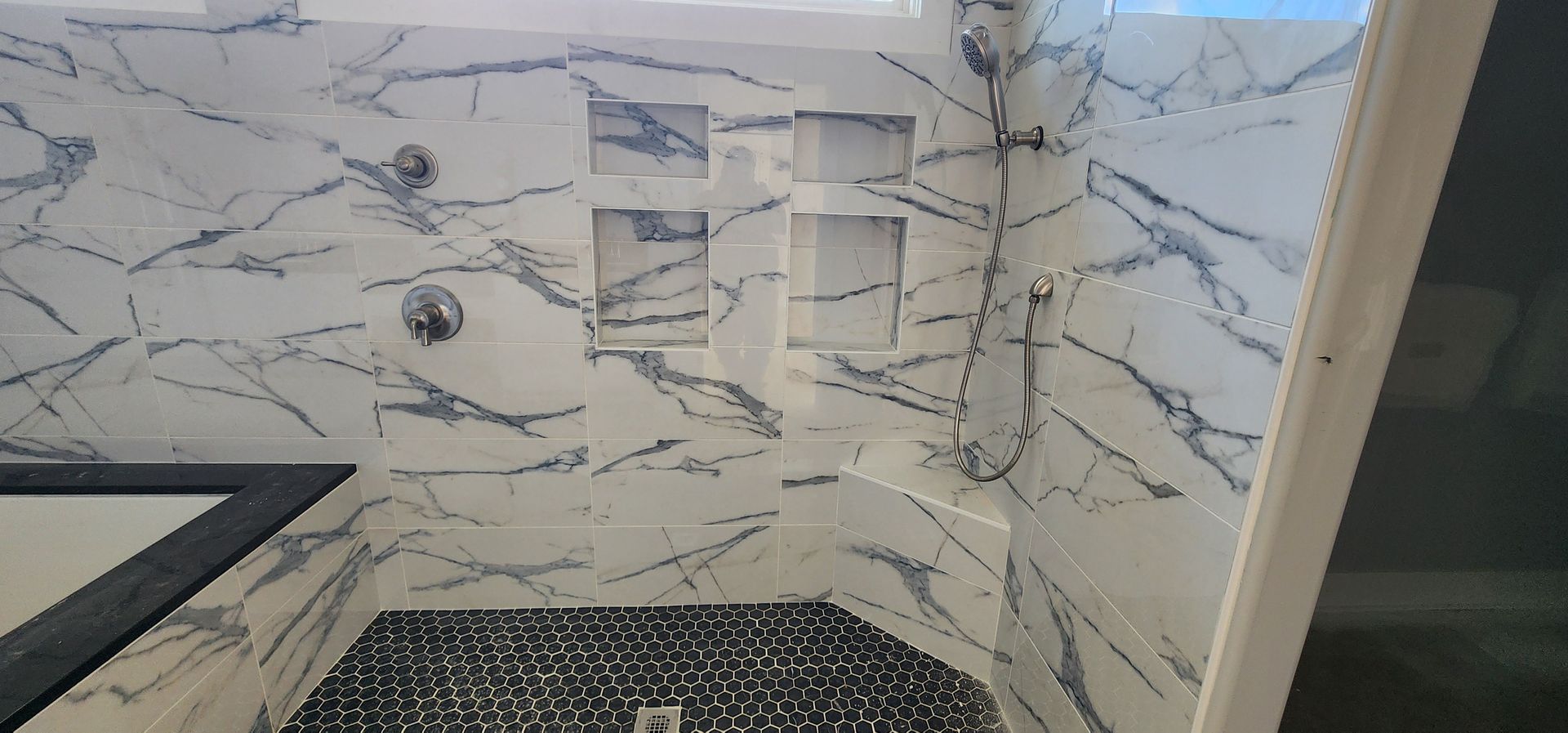 A modern shower with white marble-like tile, built-in shelves, and a dark stone floor.