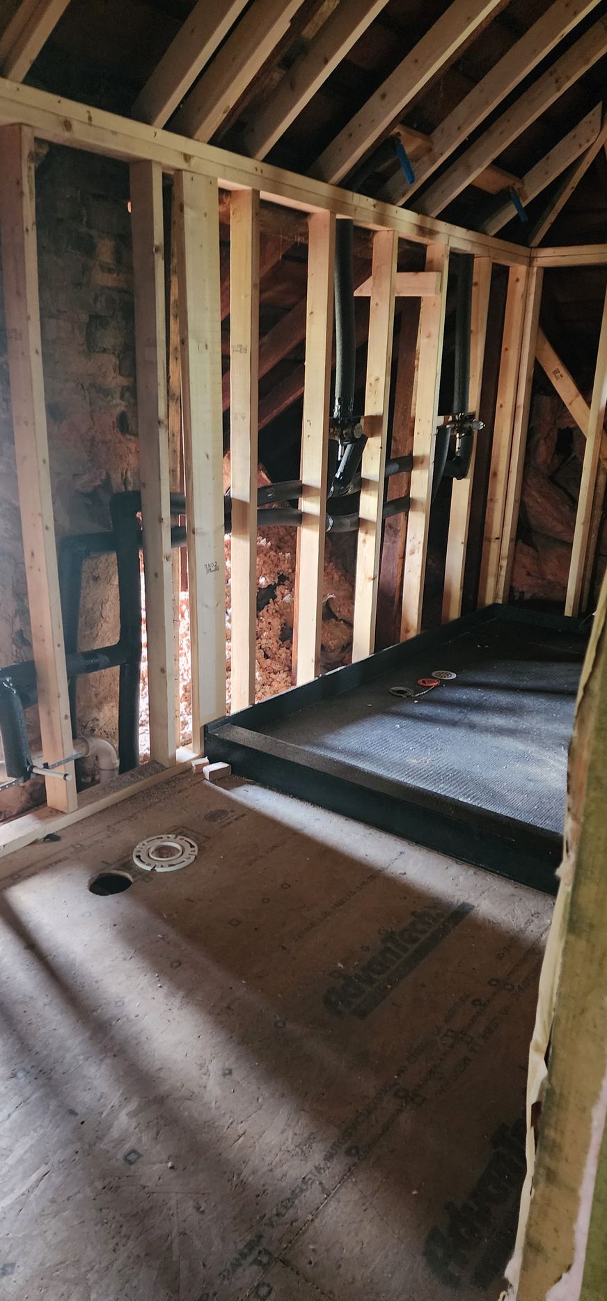 Interior framing of a bathroom with shower base installed. Studs, rafters, and dark plumbing pipes are visible.