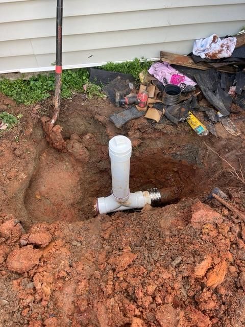 Excavation site with white PVC pipes in the ground, a shovel, and debris alongside a house.