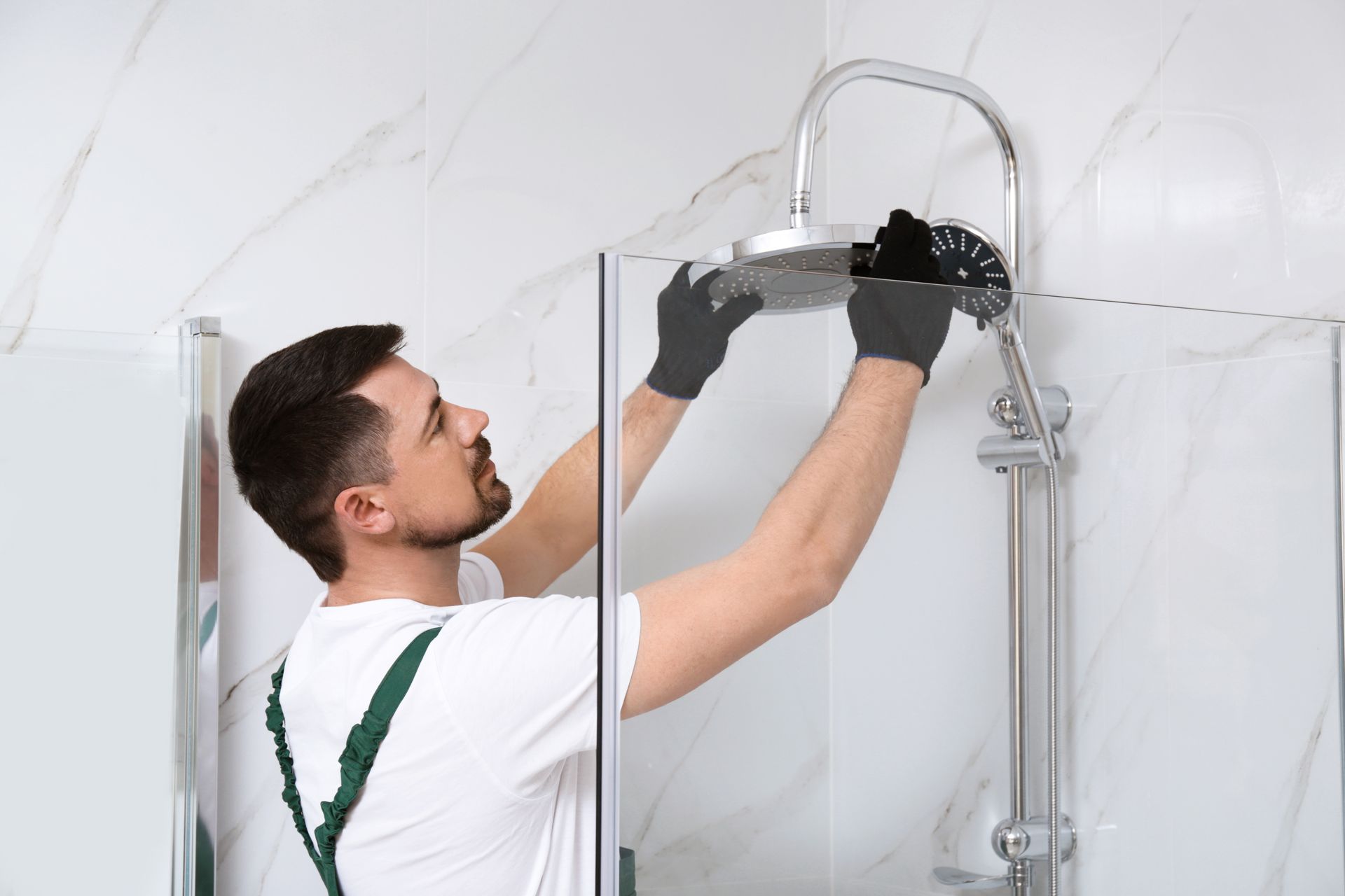 plumbers in cleveland nc Man in white shirt and green overalls installing a shower head in a bathroom, wearing gloves.