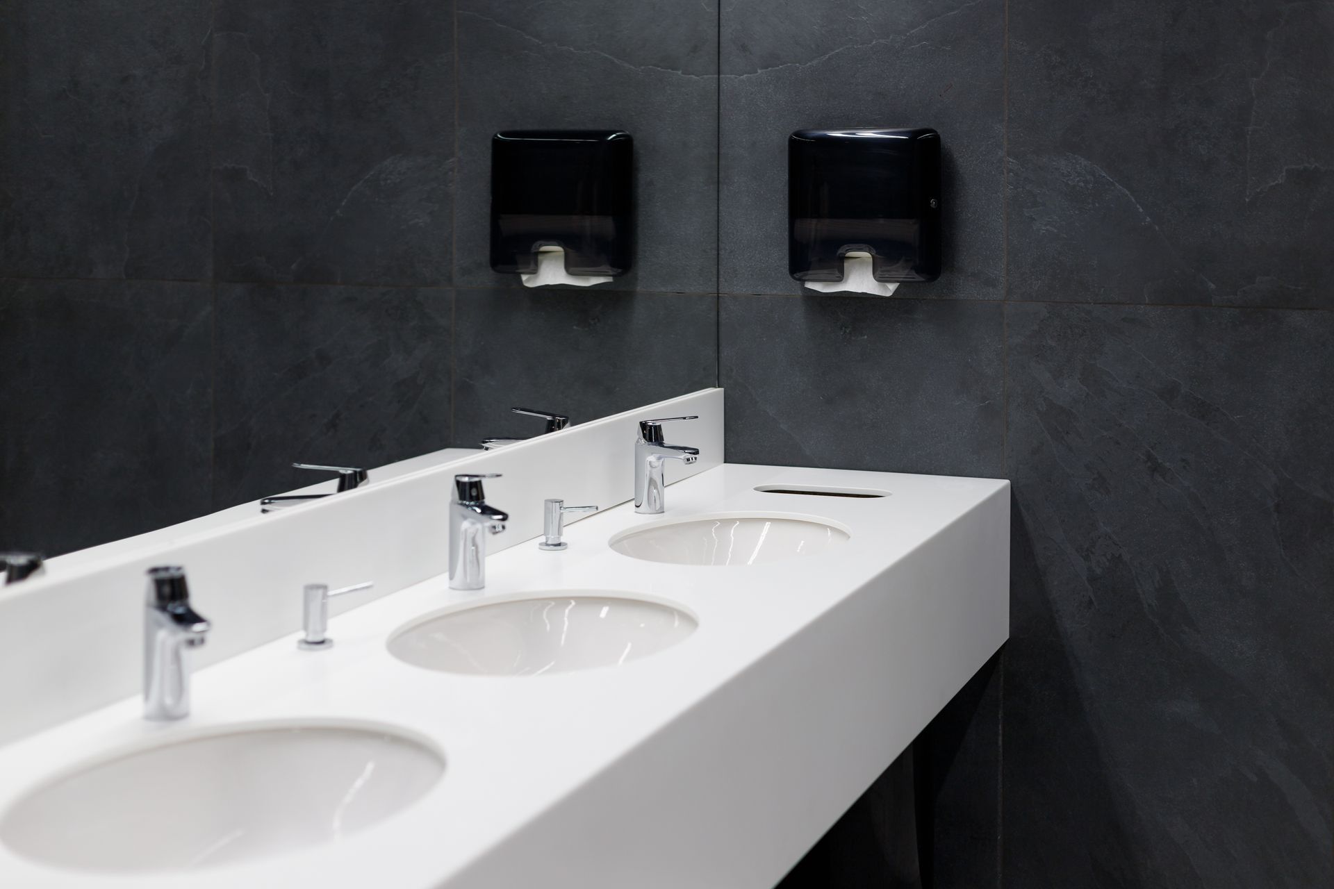 White sinks and silver faucets in a modern restroom with black dispensers against a dark tiled wall.
