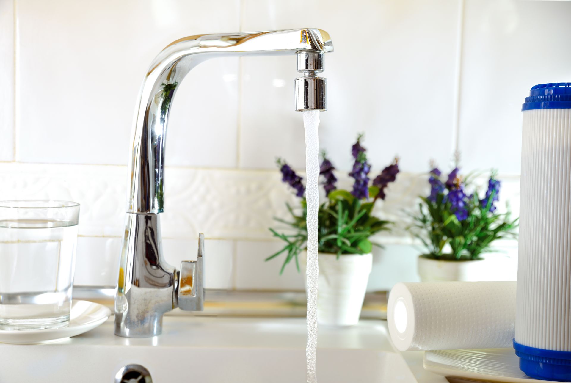 A chrome faucet dispensing water into a sink, with a glass of water, filter, and flowers.