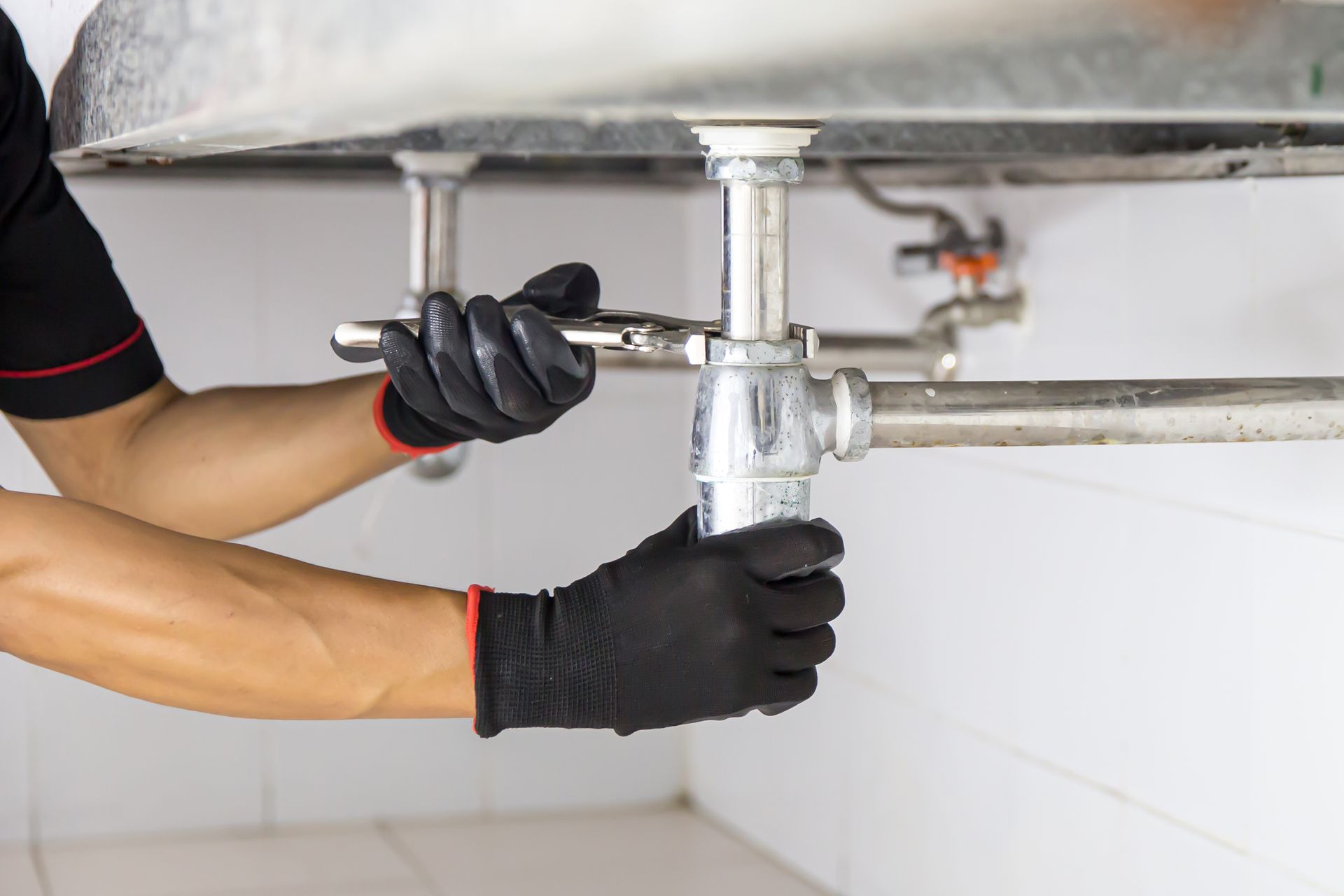 drain cleaning cleveland nc Plumber in black gloves using a wrench to work on pipes under a sink.