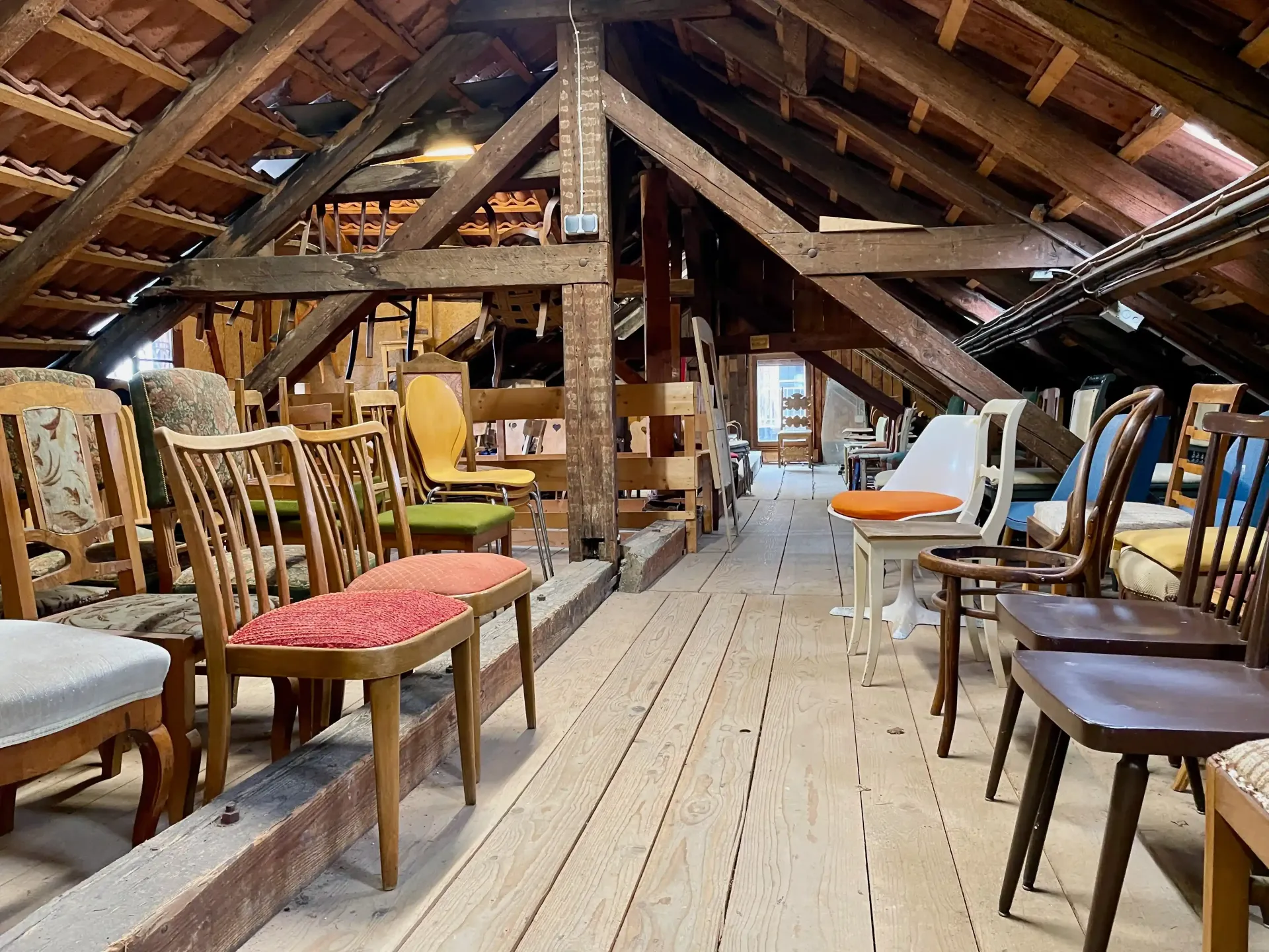 Attic room with rows of assorted chairs and wooden beams, viewed down a central aisle