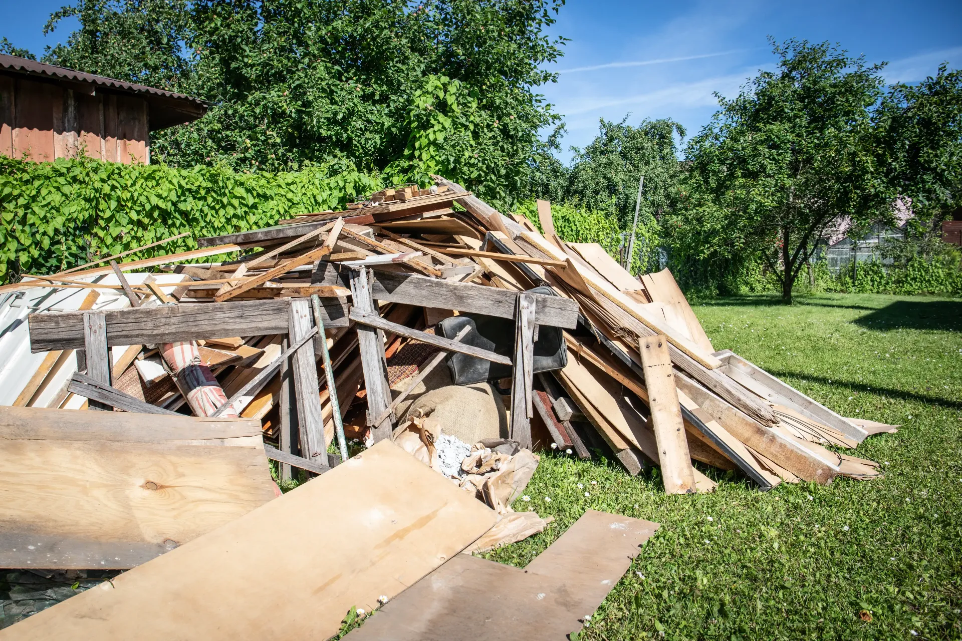 Collapsed wooden structure in a grassy yard under bright sunlight, with trees and a shed nearby