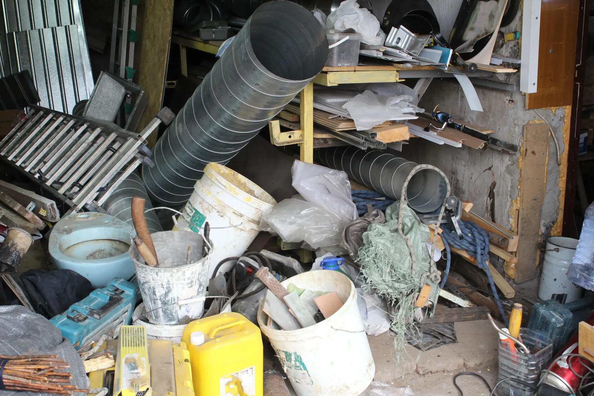 Cluttered storage area with buckets, tools, and debris piled haphazardly around a black duct pipe.