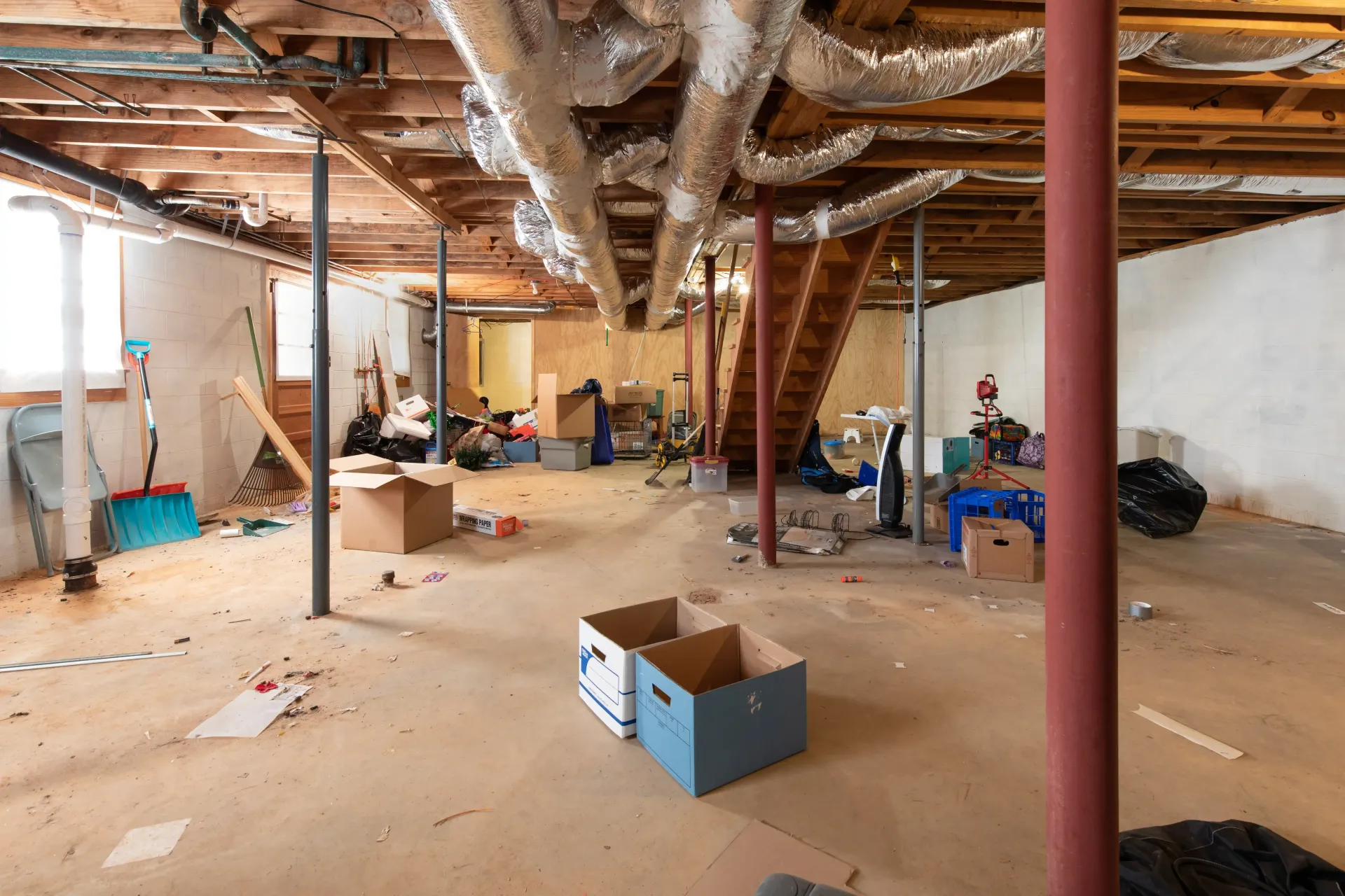 Unfinished basement with exposed beams, pipes, scattered boxes, and construction debris on the floor