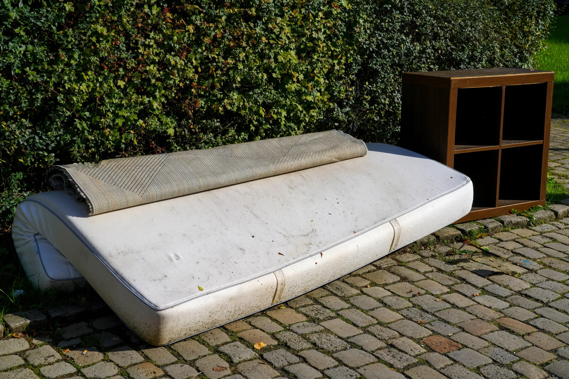 Mattress and brown shelf outside on a brick path.