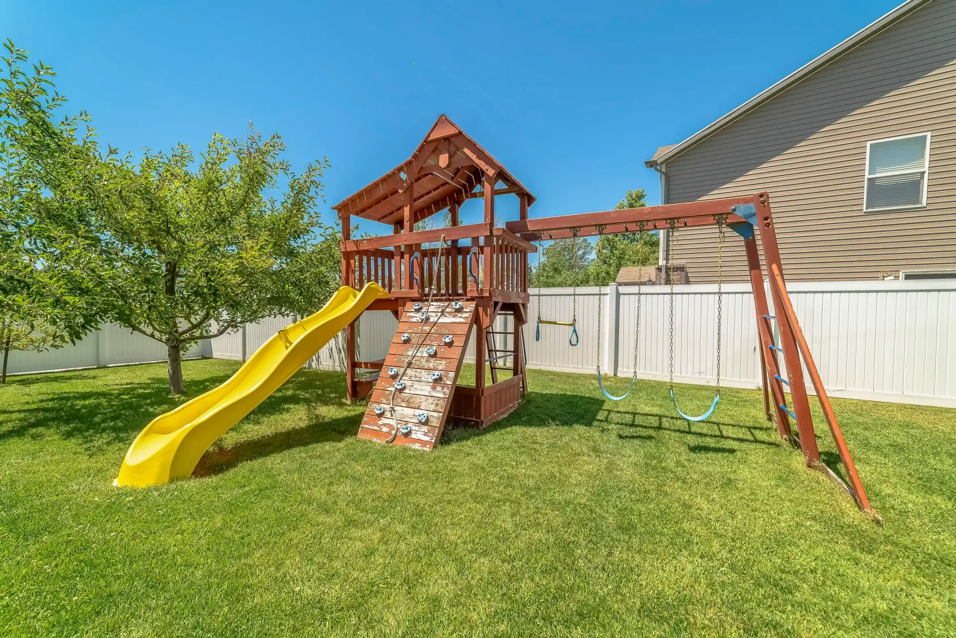 Backyard wooden playset with yellow slide, climbing wall, and swing set beside a white fence.