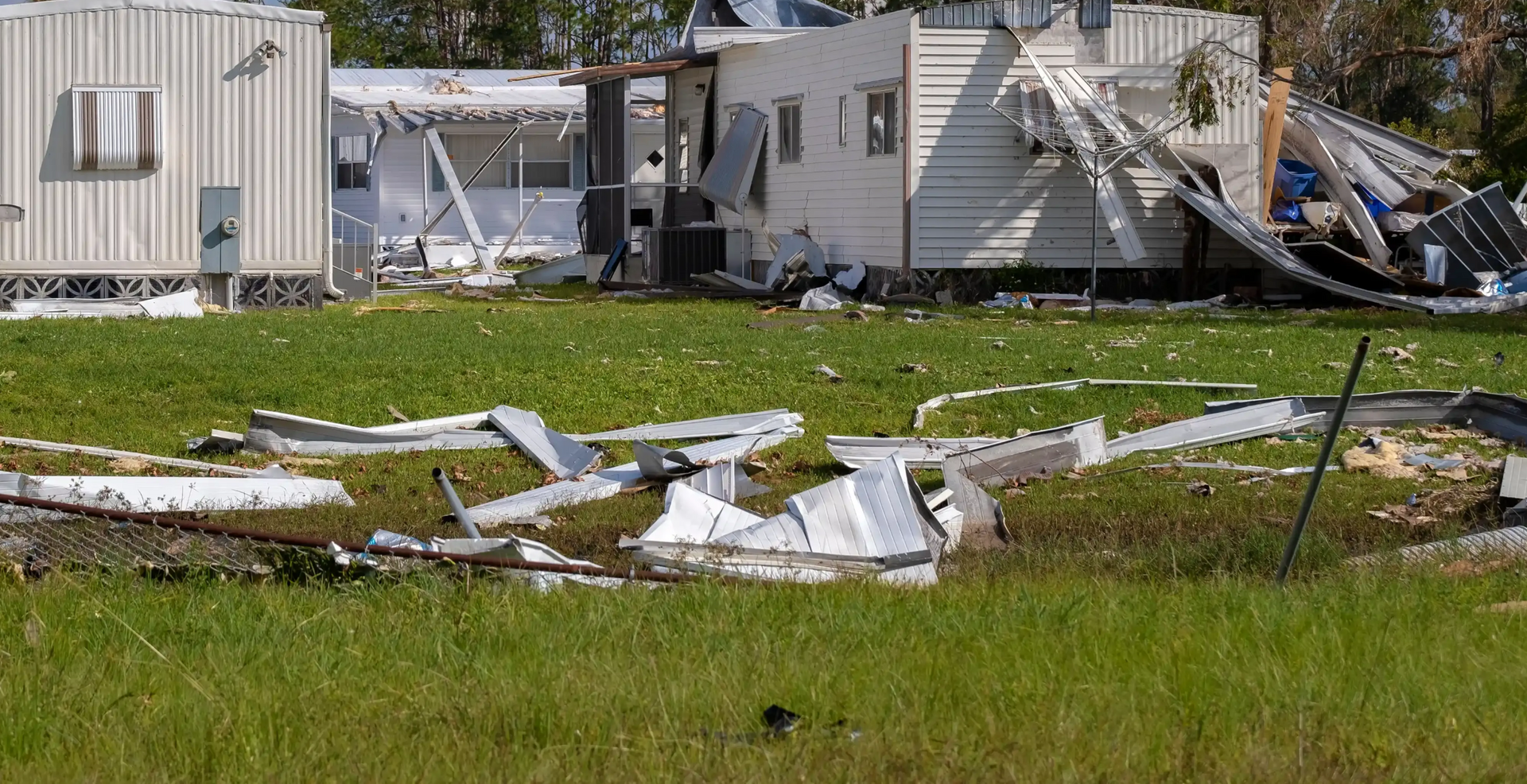 Damaged white trailer homes amid storm debris under a partly cloudy sky