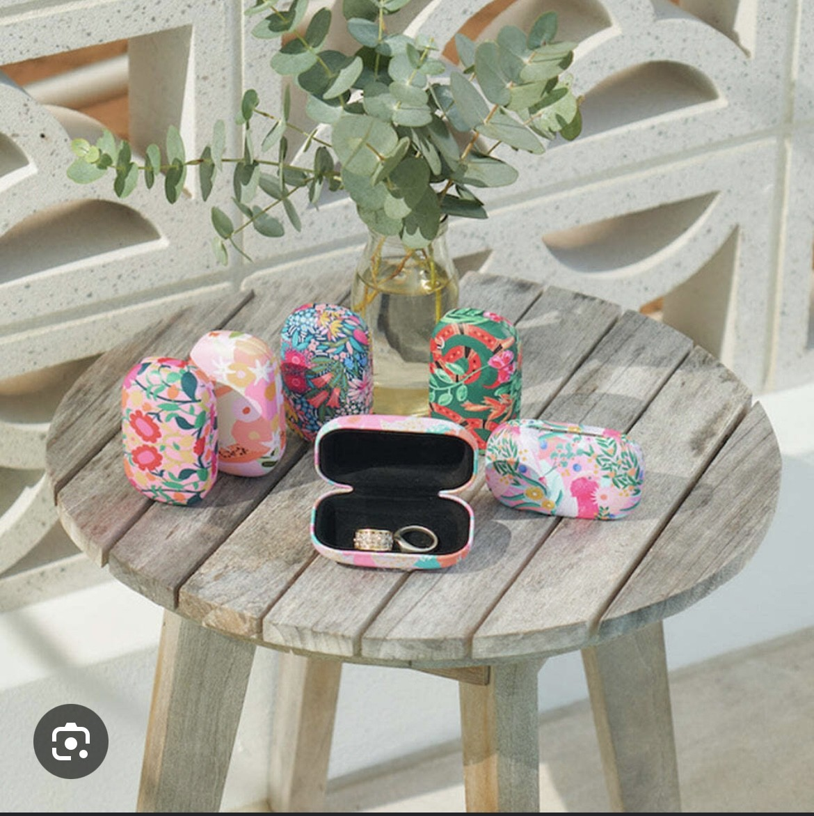 Floral patterned eyeglass cases on a rustic wooden stool with a small vase of greenery.