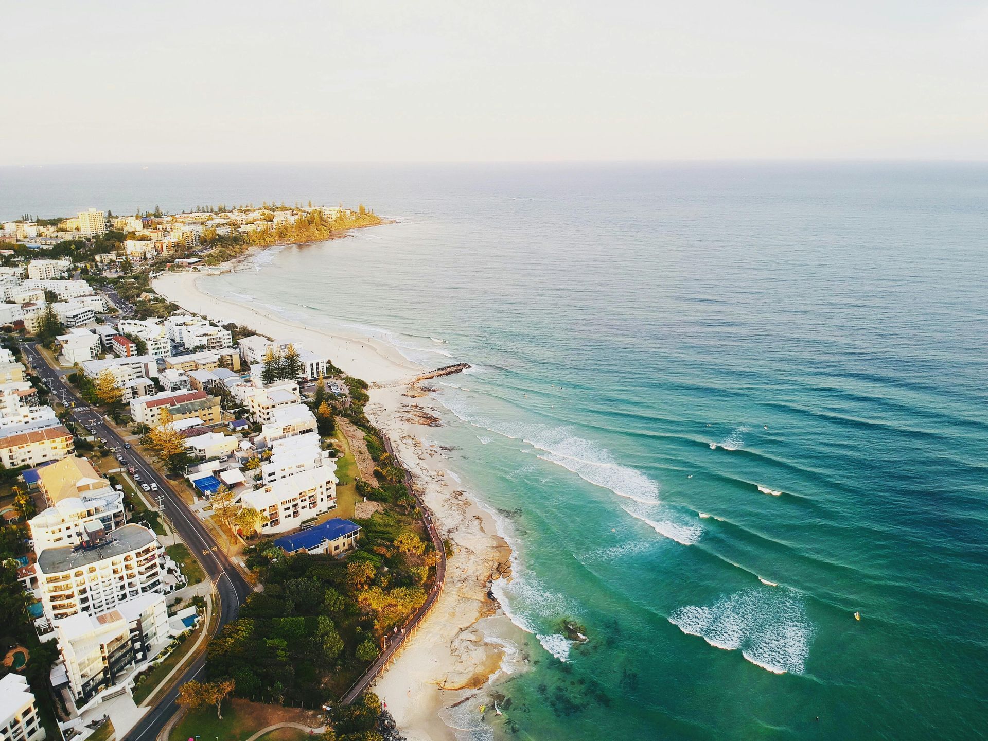Aerial view of a coastal town with white buildings along a sandy beach and turquoise ocean waves.