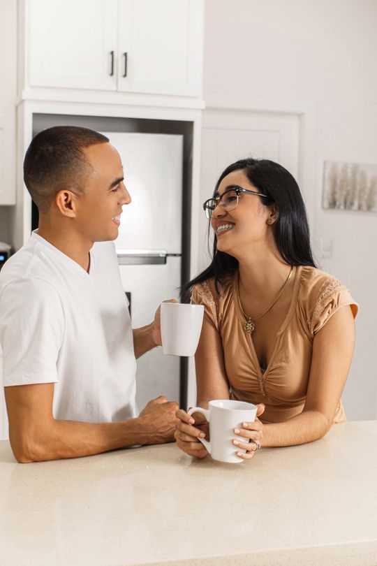 Couple in kitchen smiling, holding mugs, looking at each other, near a refrigerator and cabinets.