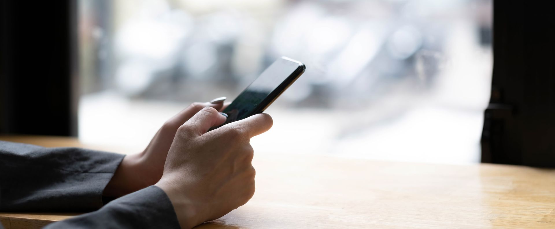 Hands holding a smartphone, tapping and swiping. Background is blurred with light shining through a window.