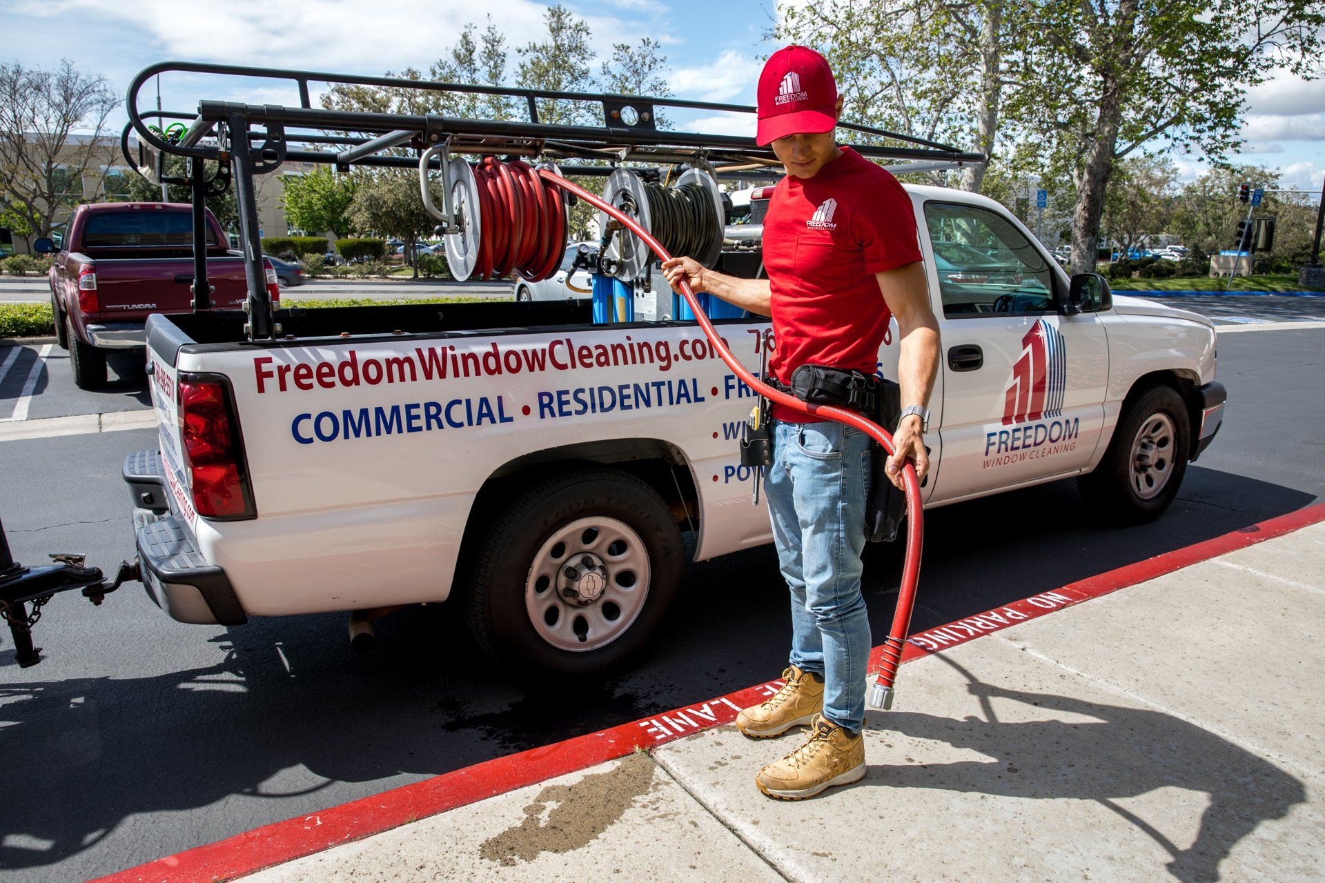 Window cleaner in red shirt and cap stands by a white commercial van with hose reel and ladder rack.