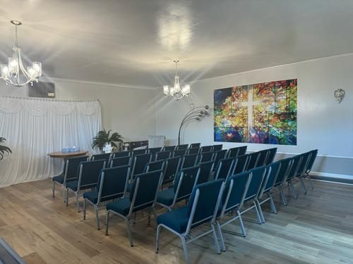 Empty ceremony room with rows of blue chairs, a colorful cross painting, and chandeliers.