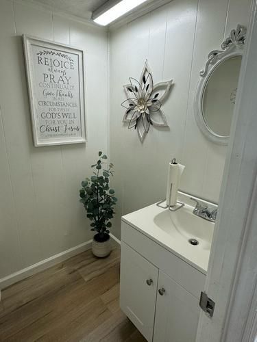 Small, white-walled bathroom with a sink, mirror, and decorative flower. Includes a plant and sign.