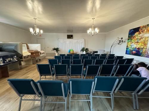 Funeral home interior with rows of blue chairs, two open caskets, and a stained glass window.