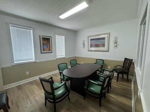 Conference room with oval table, green chairs, art, windows with blinds, and wood-look flooring.