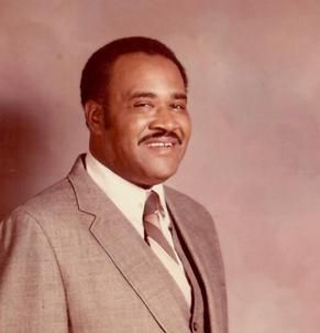 Man in a suit smiles at the camera, posing against a muted background.