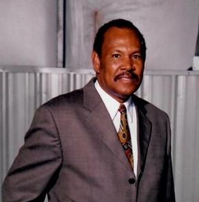 Man in a suit smiles, standing indoors near industrial equipment.