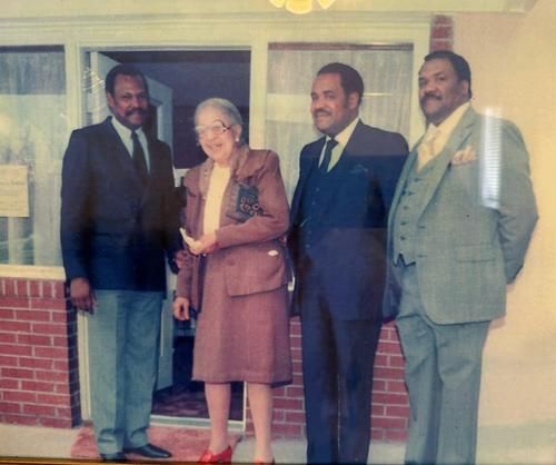 Four formally dressed people pose outside a building. Older woman in a suit stands with three men in suits.