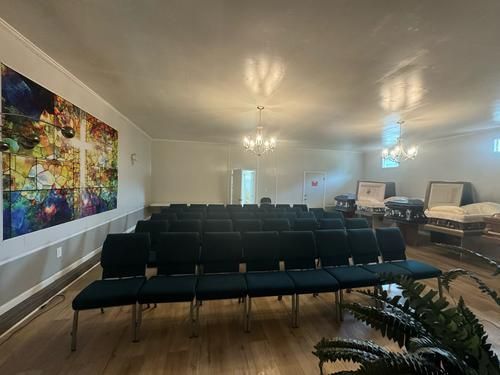 Rows of dark chairs face a room with a colorful painting on the wall, chandeliers, and potted plant.