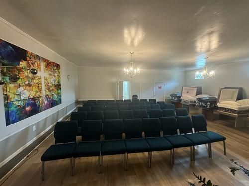 Interior of a funeral home chapel with rows of chairs, a colorful wall art, and caskets in the background.