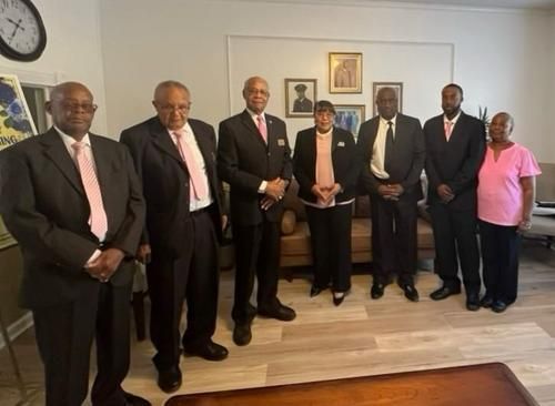 Group of people in formal attire pose inside, likely for a photo; men wear black suits, pink ties.