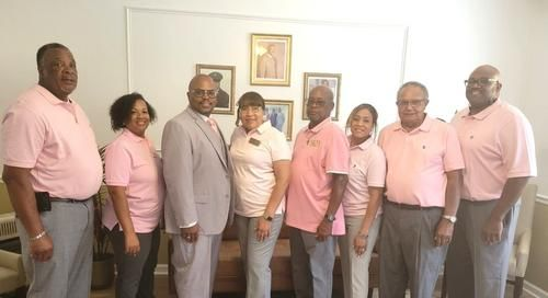 Group of people in pink shirts and gray pants standing indoors, posing.