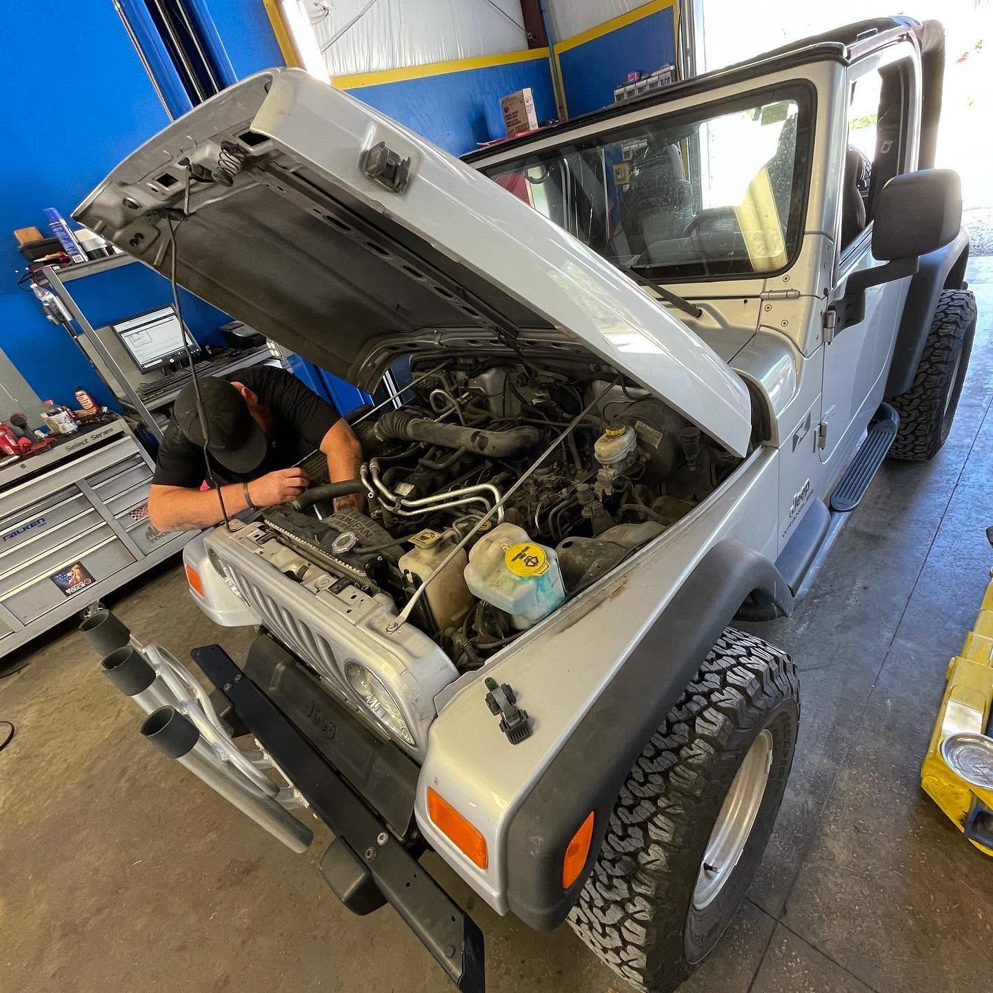 Mechanic working under the hood of a gray Jeep in an auto repair shop